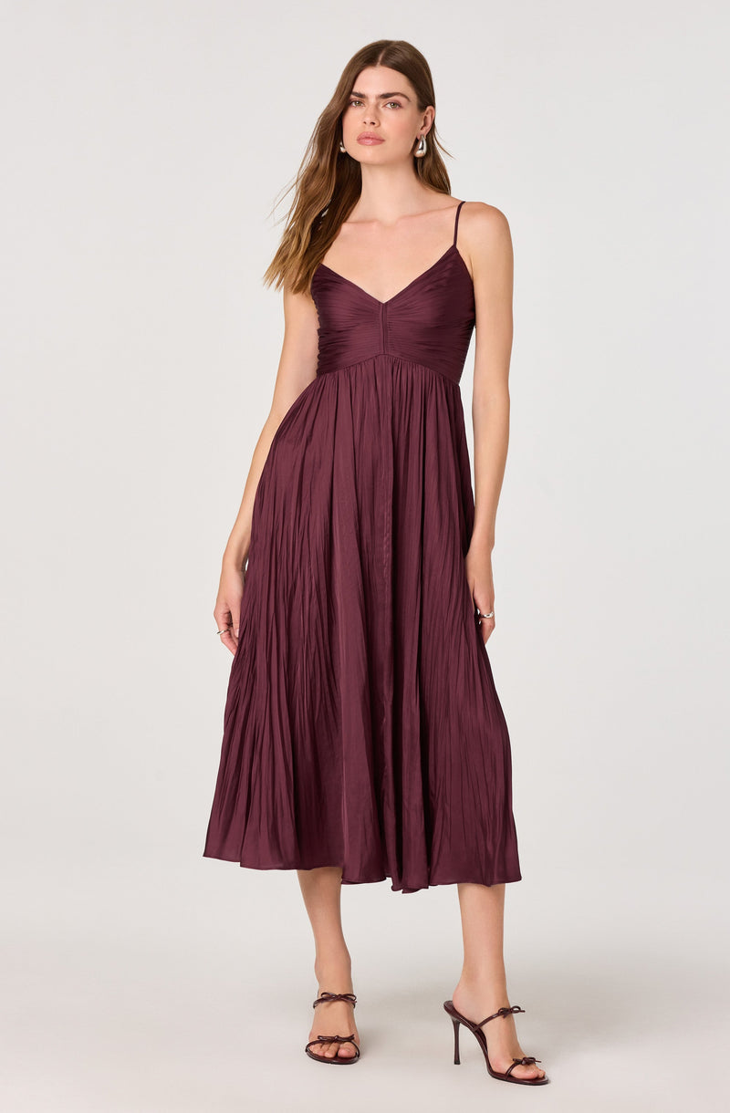 A woman is wearing a sleeveless, deep burgundy dress with thin spaghetti straps. The bodice is ruched and fitted, while the skirt is long, flowing, and pleated, reaching mid-calf. She is also wearing high-heeled sandals with ankle straps.