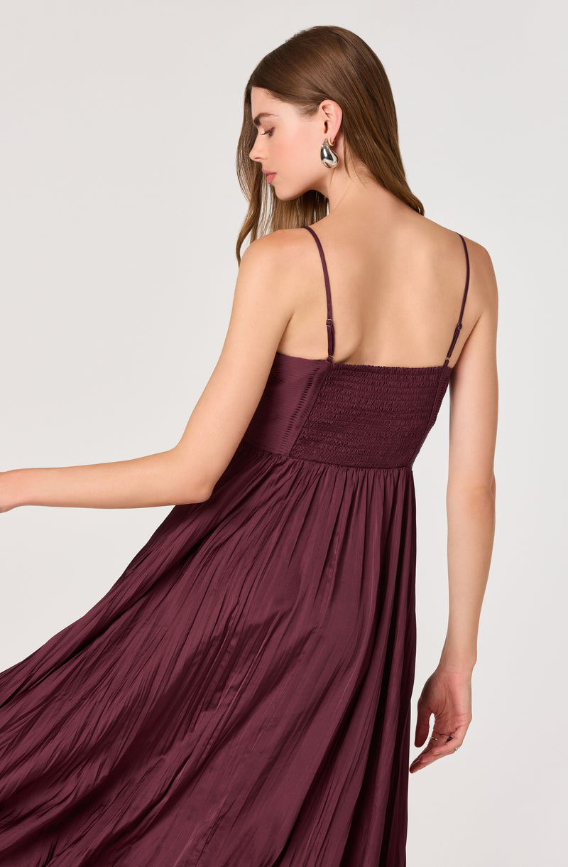 A woman is wearing a sleeveless, deep burgundy dress with thin spaghetti straps. The dress has a fitted, ruched bodice and a flowing, pleated skirt made of shiny satin-like fabric. She has long, straight brown hair and is accessorized with large, silver hoop earrings.