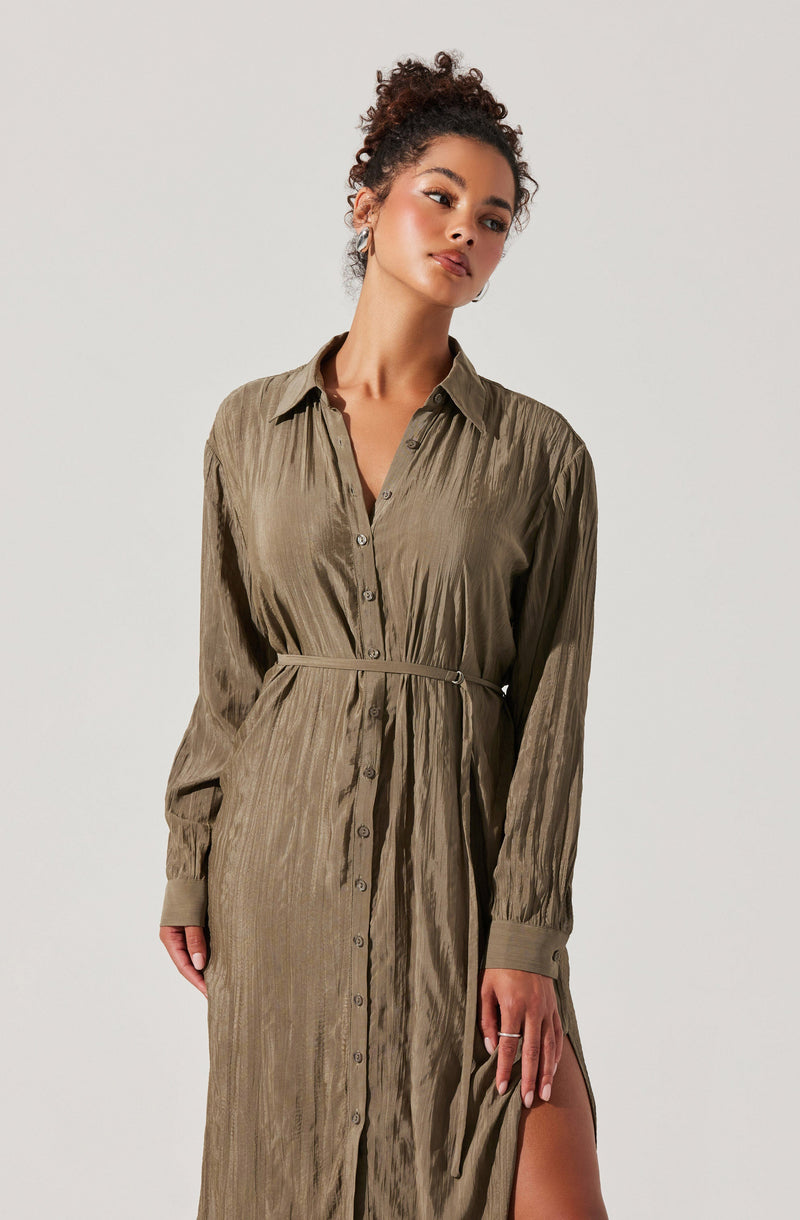 A woman is wearing a long, button-up dress made of shiny, textured fabric in an olive green color. The dress features a collar, long sleeves with cuffs, and a matching belt that cinches at the waist. The dress also has a high slit on one side, revealing part of her leg.