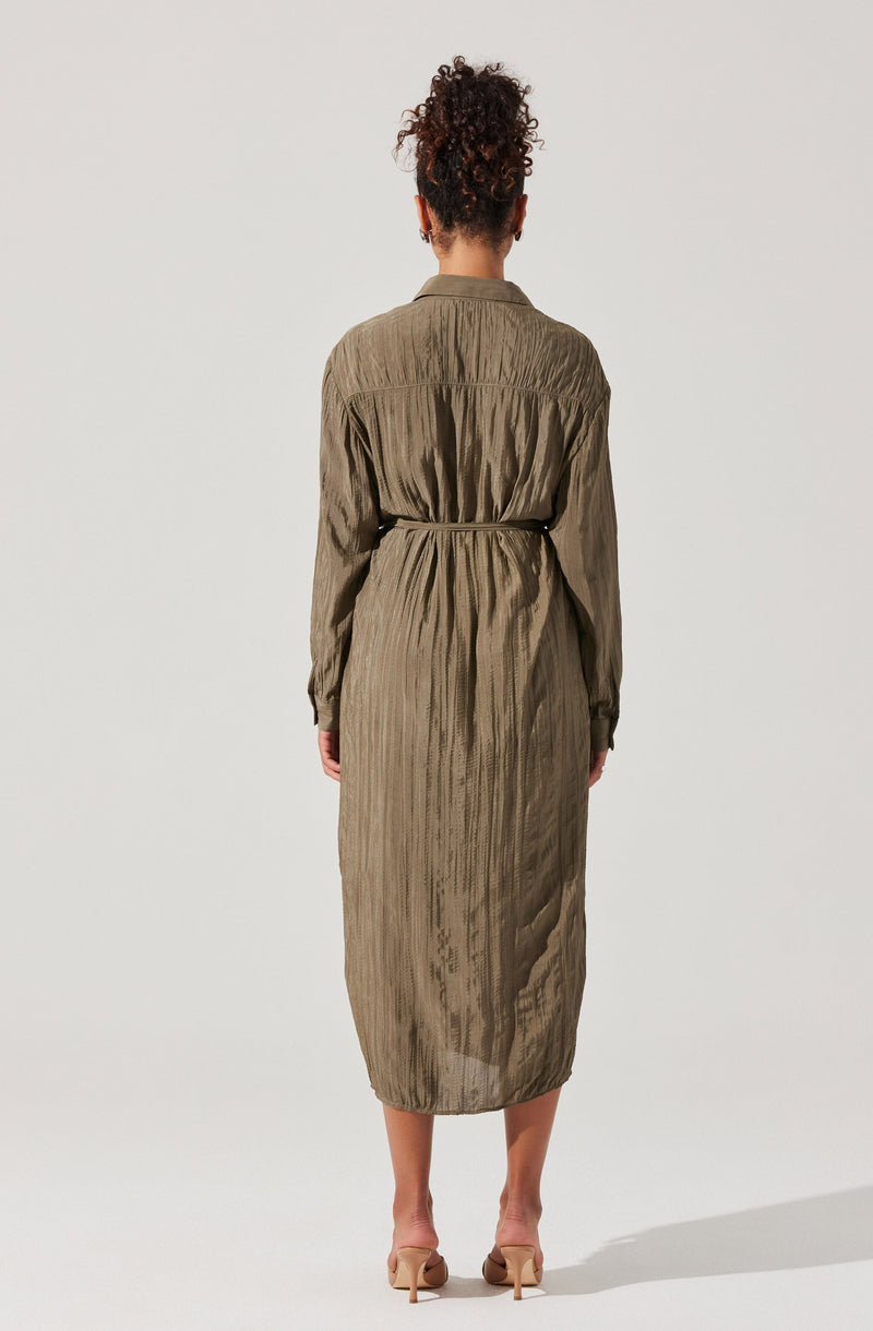 The woman is wearing a long, olive-green dress with a subtle vertical stripe pattern. The dress has long sleeves, a collar, and a waist belt made of the same fabric, which cinches the waist. The material appears lightweight and slightly textured. She is also wearing nude-colored high-heeled shoes.