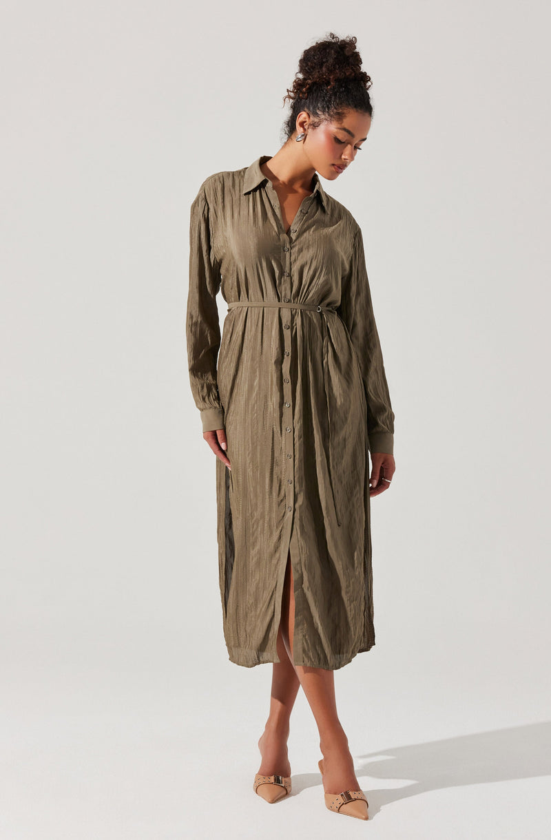 The dress is a long, button-down shirt dress in an olive-green color, featuring a pointed collar, long sleeves with buttoned cuffs, and a drawstring at the waist. It has a front slit starting from the bottom hem, giving it a relaxed yet elegant appearance. The fabric appears to have a slight sheen and crinkled texture. The woman is also wearing nude-colored pointed-toe heels with ankle straps and buckle details.