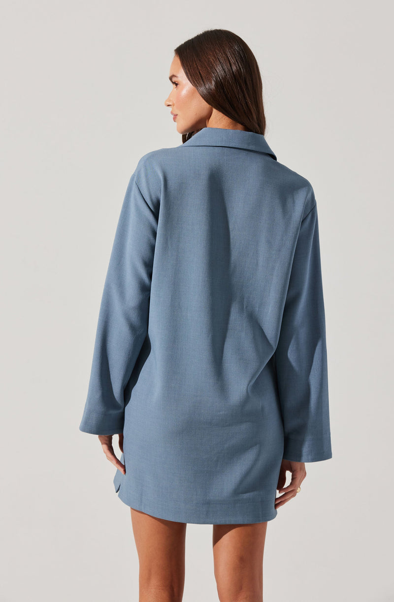 A woman is wearing a light blue, loose-fitting shirt dress with long sleeves and a collar. The dress has a simple, minimal design with no visible embellishments.
