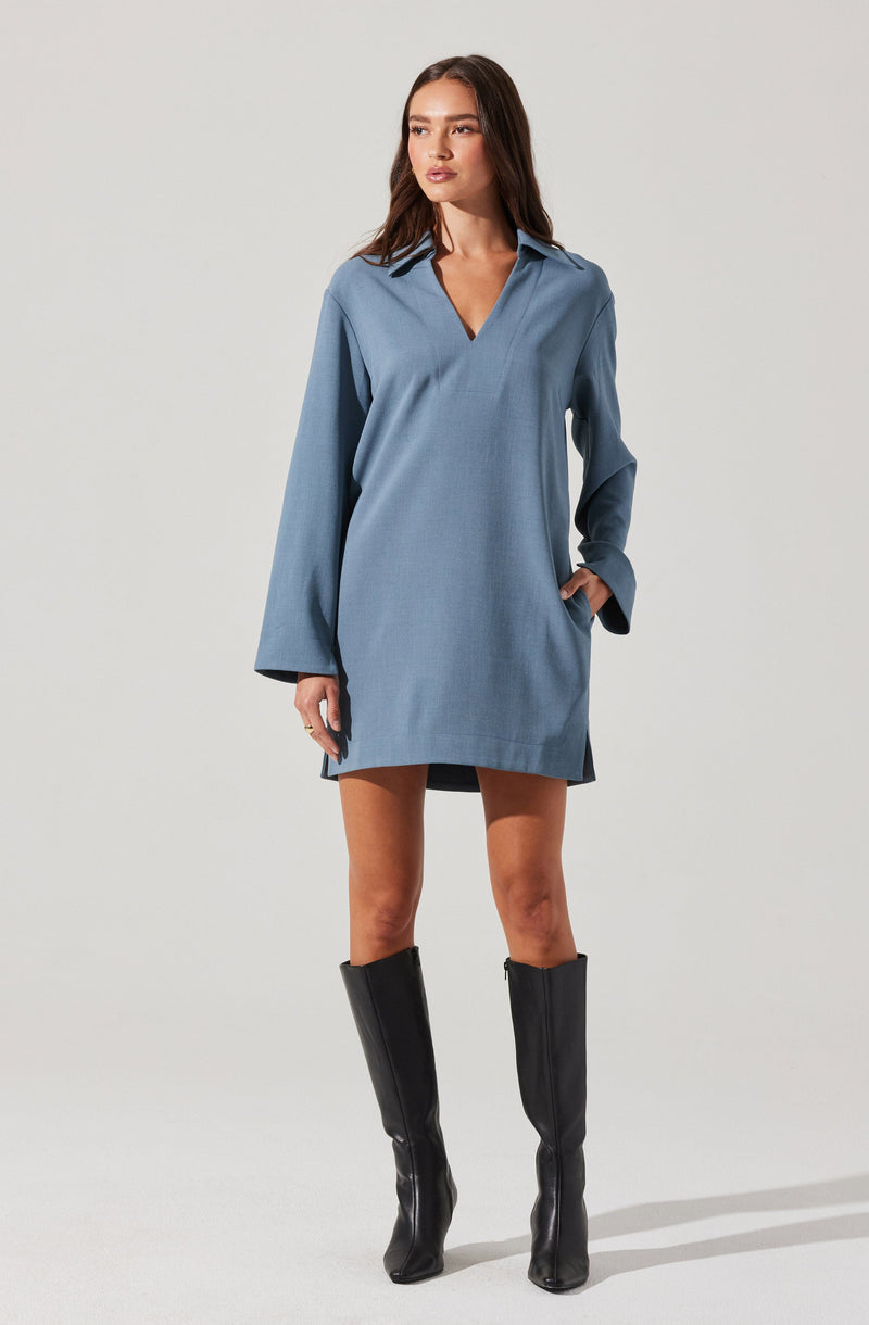 A woman is wearing a loose-fitting, light blue, long-sleeve dress with a V-neck collar. The dress has a relaxed silhouette and falls to mid-thigh length. She is also wearing knee-high black leather boots with a slight heel.