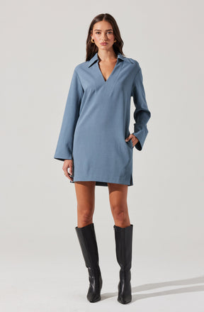 A young woman is wearing a loose-fitting, long-sleeved, light blue shirt dress with a collar and V-neck opening. The dress has a relaxed silhouette that falls above the knees. She pairs the outfit with tall black knee-high boots.