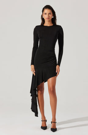 A woman is wearing a black, long-sleeved dress with a fitted bodice and a high neckline. The dress has an asymmetrical hemline with a layered ruffle detail hanging down one side, creating a dramatic and elegant look. She is also wearing black high-heeled shoes with ankle straps.