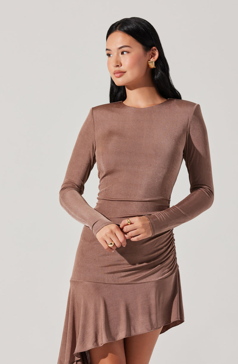A woman wearing a fitted, long-sleeved, taupe-colored dress with a round neckline. The dress features a ruched side detail on the skirt and has a slight asymmetrical hem with a ruffled edge. She has long, dark hair and is accessorized with gold earrings and rings.