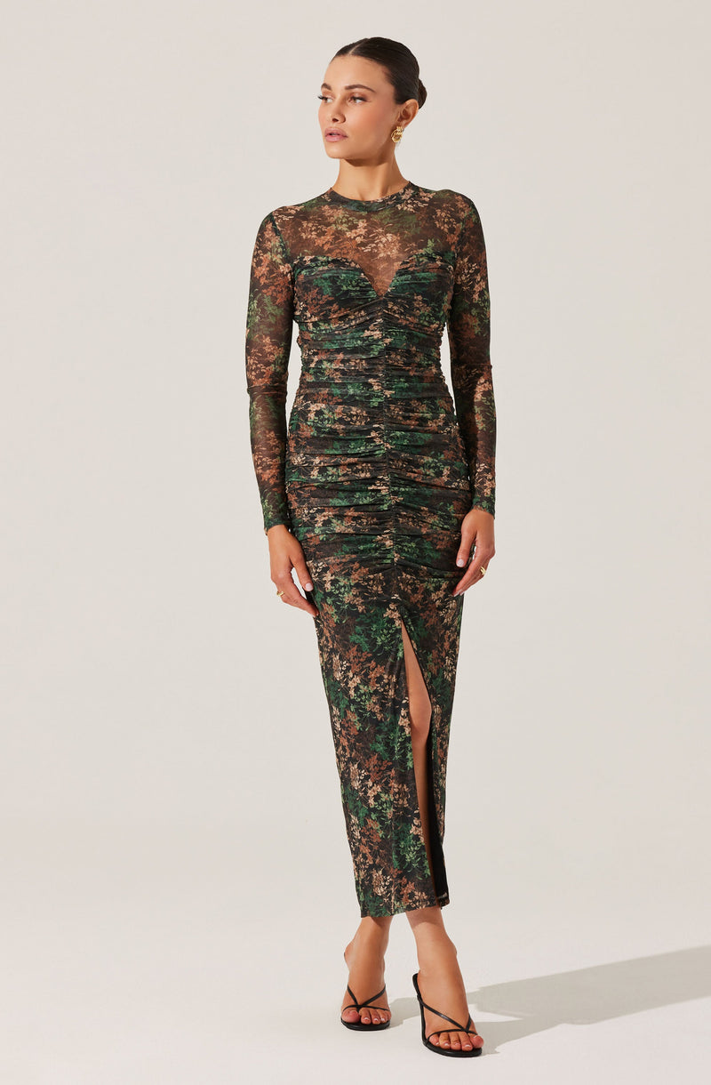 A woman is wearing a long, form-fitting dress with a camouflage pattern in shades of green, brown, and black. The dress features long, sheer sleeves and a high neckline. It has ruching along the front and a high slit at the front, revealing her legs. She is also wearing black, open-toe high-heeled sandals.
