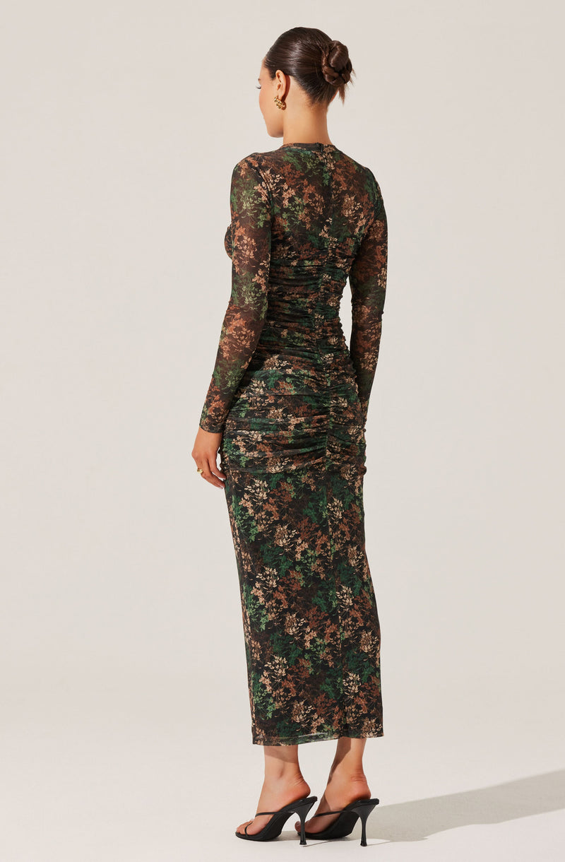 A woman is wearing a long, fitted dress made of sheer, floral lace fabric in shades of black, brown, green, and beige. The dress has long, semi-transparent sleeves and a high neckline, with a ruched design along the back that accentuates the waist and hips. She is also wearing black high-heeled sandals with thin straps.