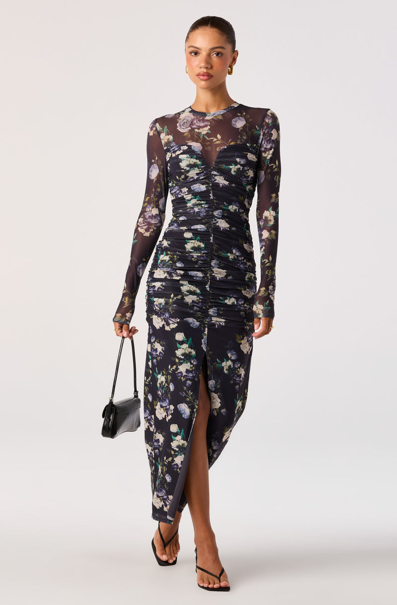 She wears a long black floral dress with sheer long sleeves, a high slit, and ruched detailing.