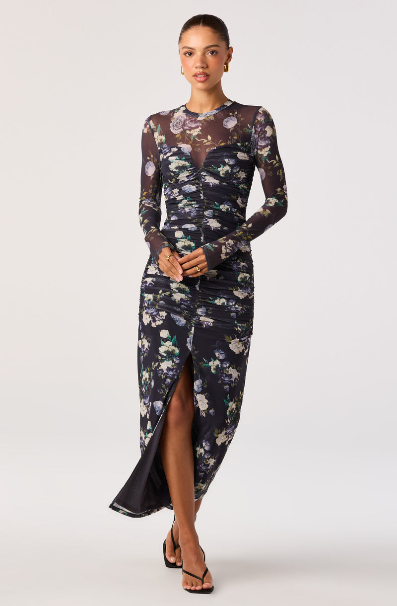 Girl wearing a long, black floral dress with sheer long sleeves and a front slit.