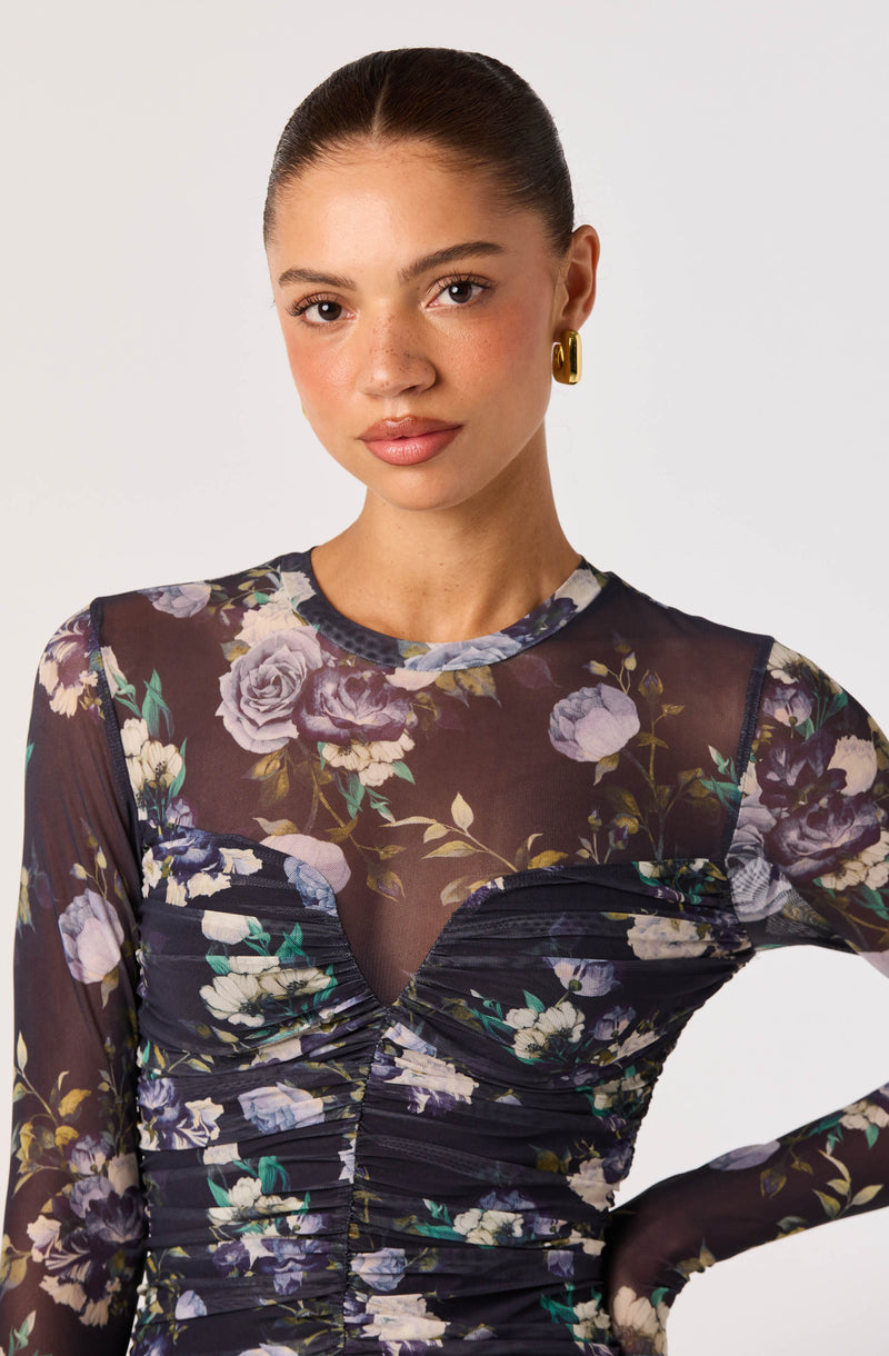 Woman in a black sheer floral long-sleeve dress with ruched details and gold hoop earrings.