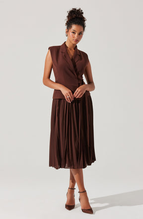 A woman is wearing a sleeveless, brown blazer-style top with a notched collar and a matching pleated midi skirt. She has on pointed-toe, ankle-strap heels that are also brown.