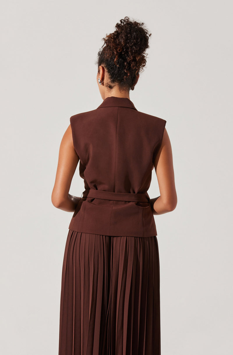 The woman is wearing a sleeveless, brown vest with a collar and a matching waist belt. She is also wearing a long, pleated skirt in a matching brown color.