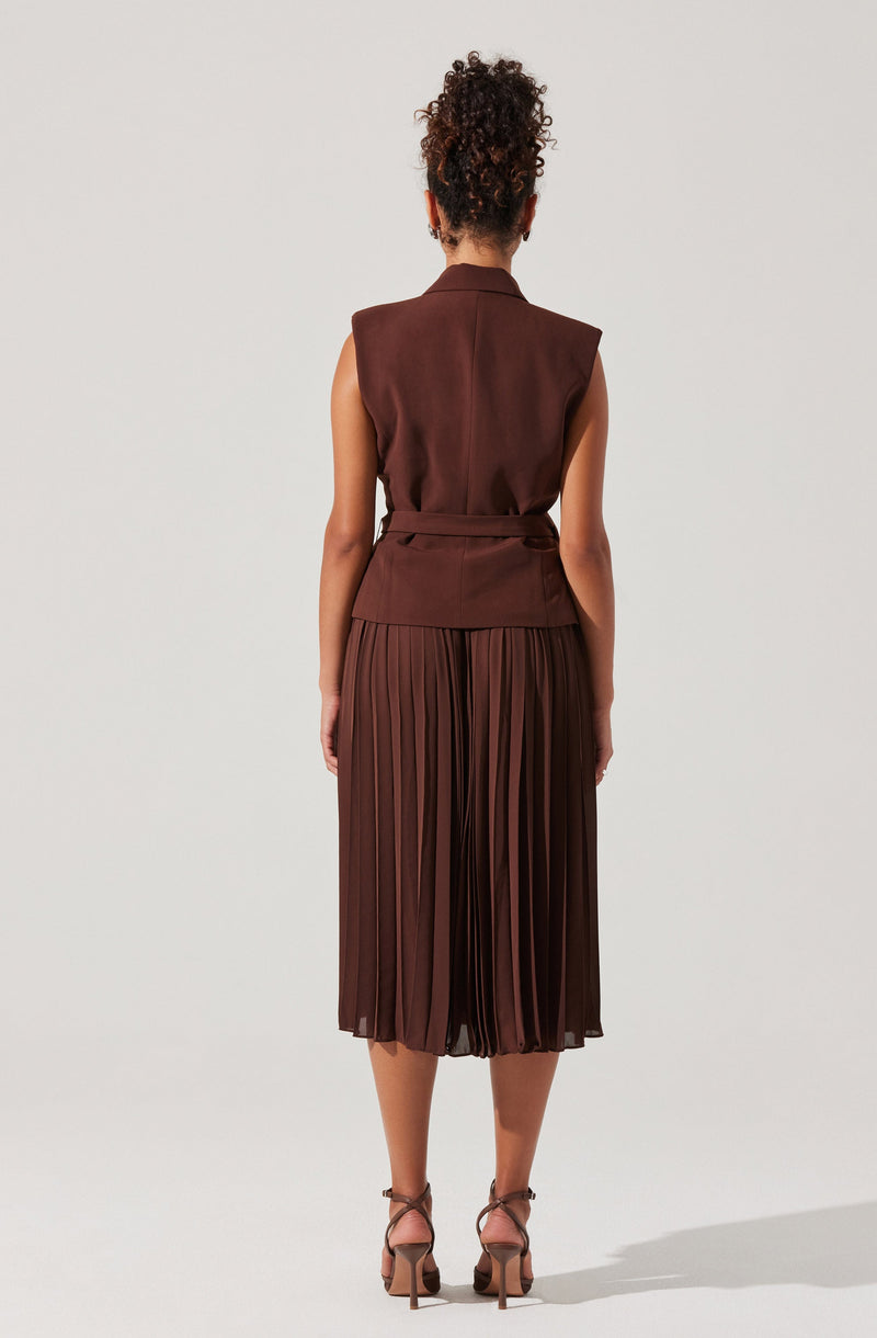 A woman is wearing a sleeveless, tailored brown vest with a belt at the waist, paired with a pleated brown midi skirt that falls below the knees. She is also wearing strappy high-heeled sandals.