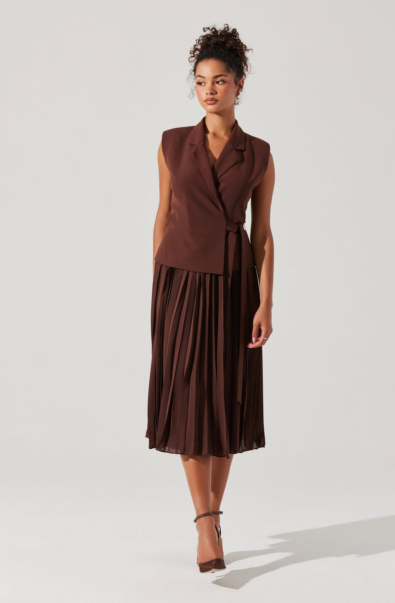 A woman is wearing a sleeveless, dark brown blazer with a tie belt and a matching pleated midi skirt. She is also wearing dark brown high-heeled shoes with ankle straps.