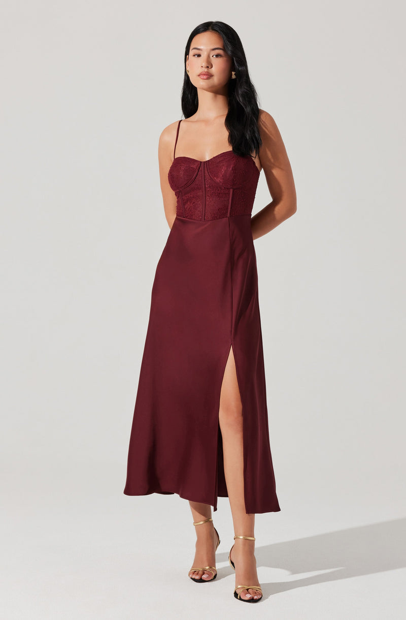 The dress is a sleeveless, spaghetti strap gown in a deep burgundy color. The bodice is fitted with a sweetheart neckline and features a lace overlay with intricate detailing. The skirt is long and flowing, with a high slit on one side that reveals the leg. The fabric appears to be satin or silk, giving it a smooth and shiny finish. The dress is paired with strappy high-heeled sandals.