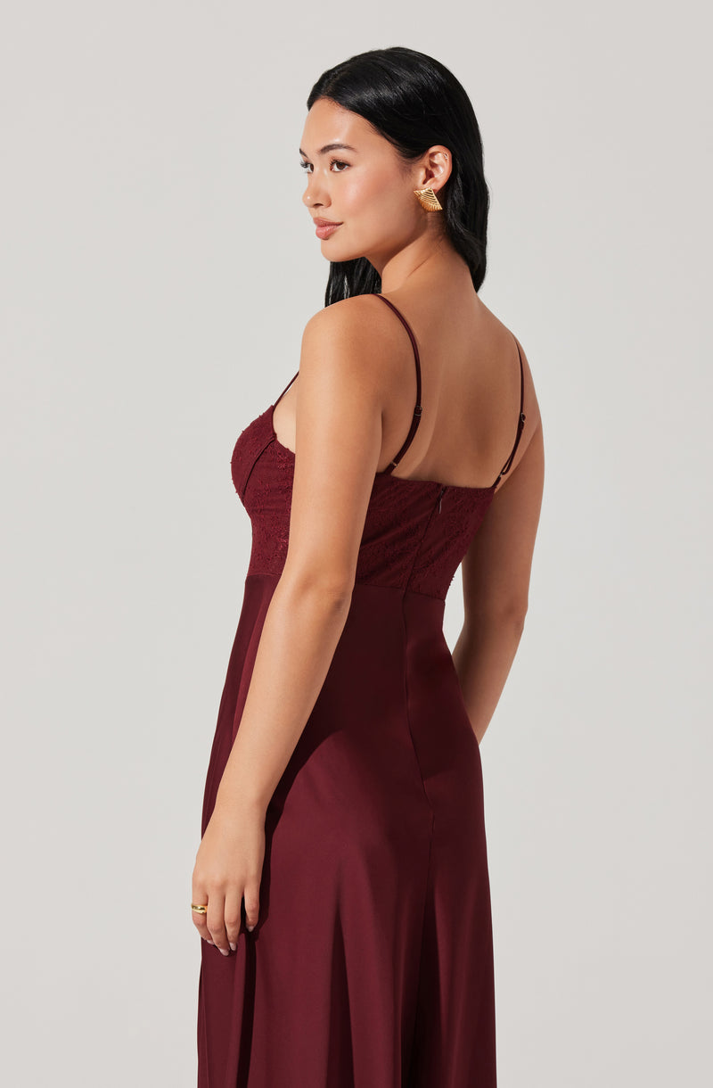 A woman is wearing a burgundy satin dress with spaghetti straps. The top of the dress features a textured, lace-like fabric, while the skirt is smooth and shiny, flowing down to the floor. She has long, dark hair and is accessorized with gold earrings and a gold ring.