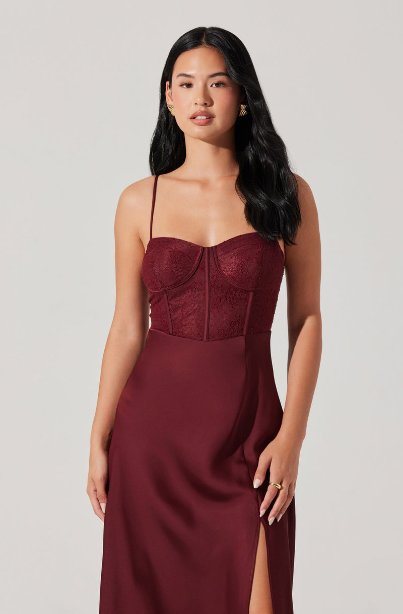 The woman is wearing a sleeveless, burgundy dress with thin spaghetti straps. The top part of the dress features a lace design with a sweetheart neckline and structured cups, while the lower part is smooth satin with a slit on one side.