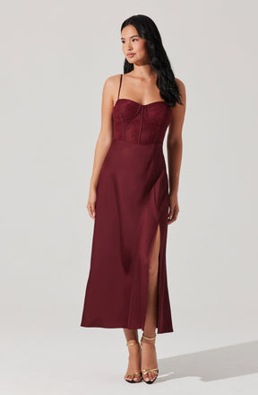 A woman is wearing a sleeveless, burgundy dress with spaghetti straps. The dress has a fitted, lace bodice with a zipper down the center and a long, flowing skirt that features a high slit on one side. She is also wearing strappy gold high-heeled sandals.
