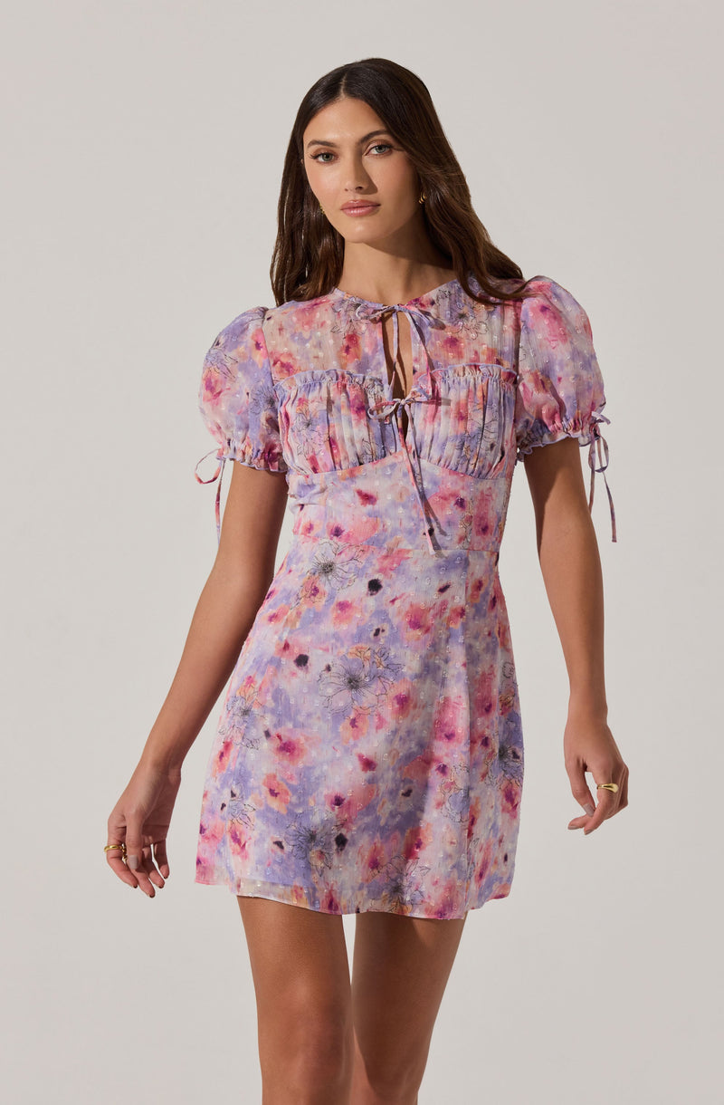 A woman is wearing a short, colorful dress with a watercolor floral pattern in shades of pink, purple, orange, and blue. The dress features puffed, short sleeves with tied bows, a round neckline with a small front opening secured by a tie, and a fitted waist.