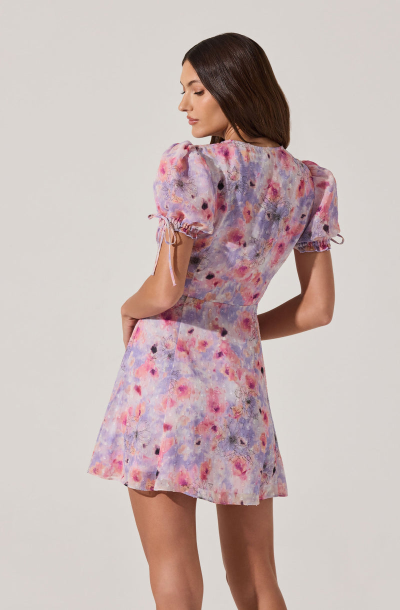 A woman is wearing a short, pastel-colored dress with an abstract, floral pattern. The dress features puffed short sleeves with small bows tied at the ends. The fabric appears lightweight and has a soft, watercolor-like print in shades of pink, purple, and blue.
