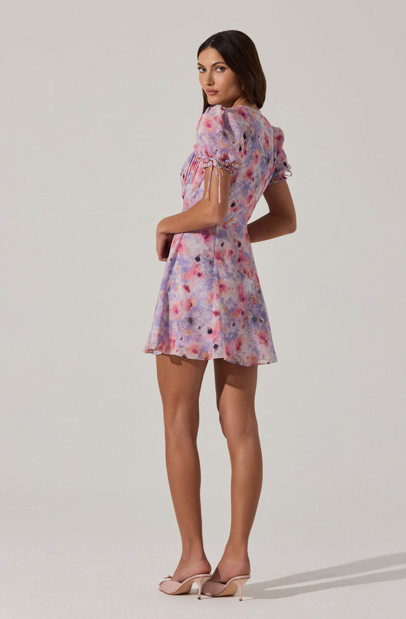A woman is wearing a short dress with puffed sleeves. The dress has a colorful, abstract pattern in shades of pink, purple, and blue. The sleeves have a delicate drawstring detail at the edges. She is also wearing light-colored high heels.