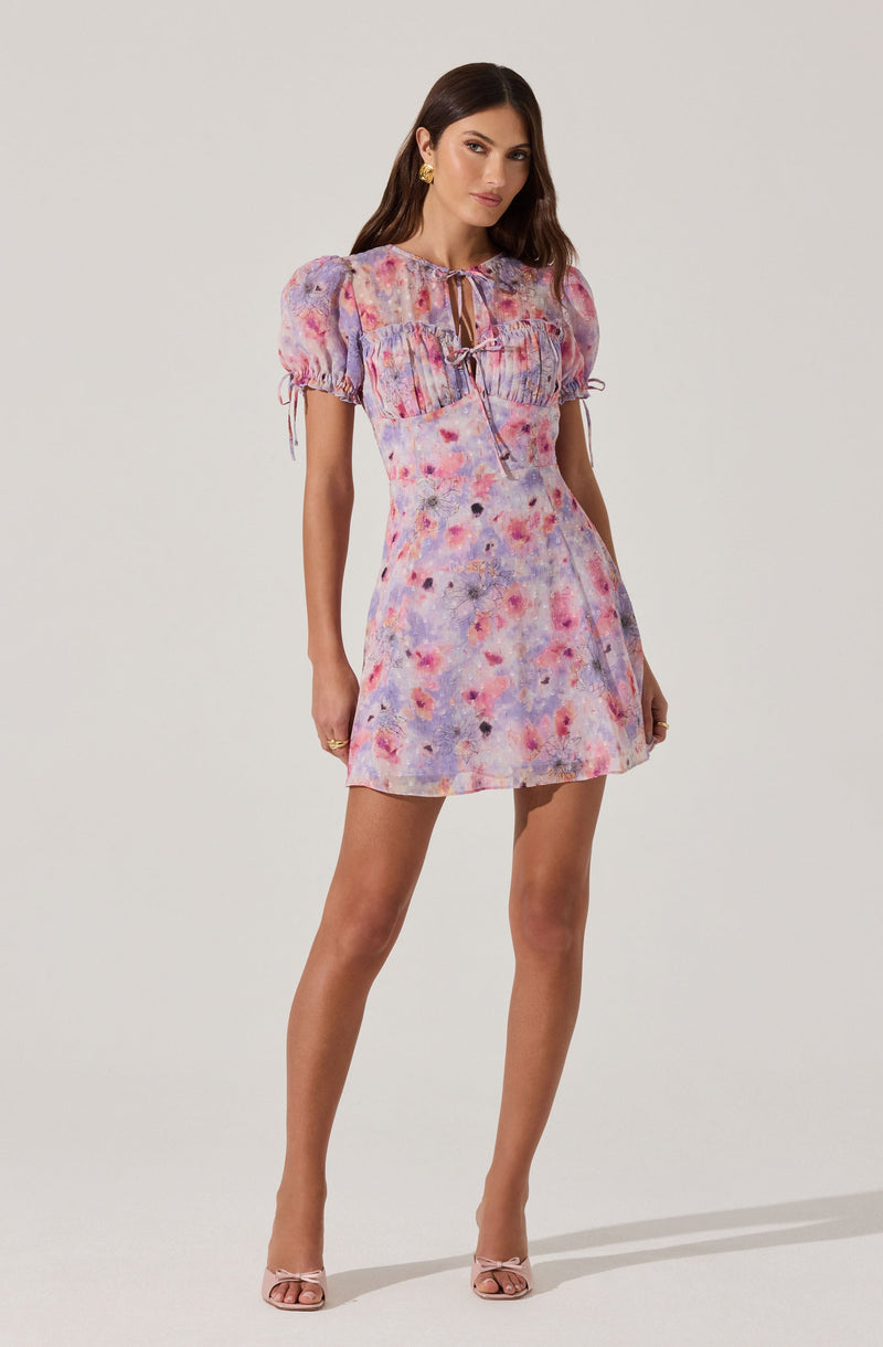 A woman is wearing a short, pastel-colored dress with a tie neckline and puffed sleeves. The dress features a watercolor-like pattern in shades of pink, purple, blue, and orange. She has on gold earrings and nude high-heeled sandals.