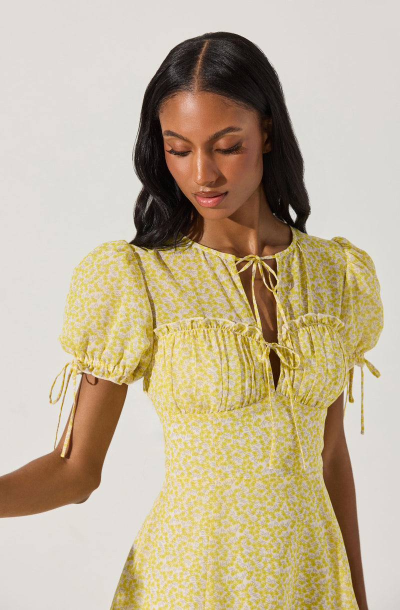 A woman is wearing a yellow dress with a white floral pattern. The dress features puffed short sleeves with ties at the edges, a ruched bust with a ruffle detail, and a tie-up neckline. The fabric appears lightweight and summery.
