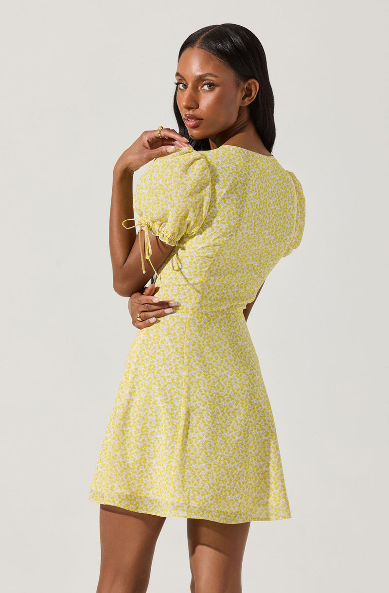 A woman is wearing a yellow dress with a subtle floral pattern. The dress features puffed short sleeves with ties at the ends, a fitted waist, and a slightly flared skirt.