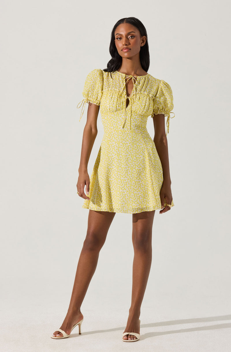A woman is wearing a yellow dress with a subtle floral pattern. The dress has puffed short sleeves with ties at the ends, a square neckline with ruffle detailing and a front tie closure. The dress has a fitted waist and a knee-length, slightly flared skirt. She pairs the dress with beige high-heeled sandals.