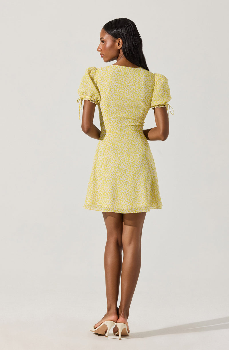 A woman is wearing a short, yellow dress with a white floral pattern. The dress has puffed short sleeves with ties at the ends, a fitted bodice, and a flared skirt. She is also wearing beige high-heeled shoes.