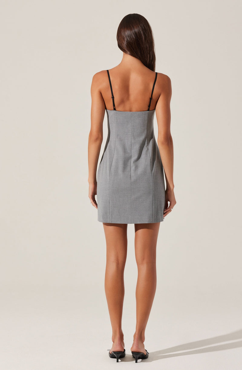 A woman is wearing a sleeveless, fitted gray dress with thin black spaghetti straps. The dress has a subtle texture and a straight hemline. She is also wearing black high-heeled shoes. Her hair is long and straight, and she is standing against a plain, light-colored background.