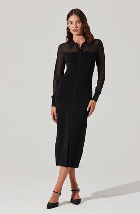 A woman is wearing a fitted, long black dress with a front zipper. The dress features sheer, long-sleeved panels on the upper chest, shoulders, and sleeves, contrasted by opaque fabric on the rest of the dress. She is also wearing black pointed-toe heels with ankle straps.