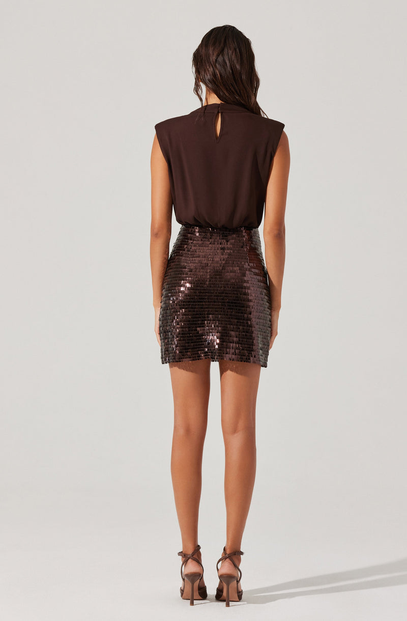A woman is wearing a sleeveless dark brown top with a small keyhole opening at the back neckline, paired with a high-waisted, short skirt covered in shiny, dark sequins. She is also wearing strappy high-heeled sandals.