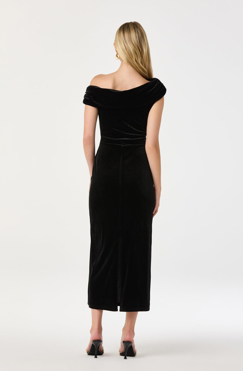 A woman is wearing an elegant, off-the-shoulder black velvet dress that is fitted through the bodice and hips, extending down to a full-length skirt with a back slit. She is also wearing black high-heeled shoes.