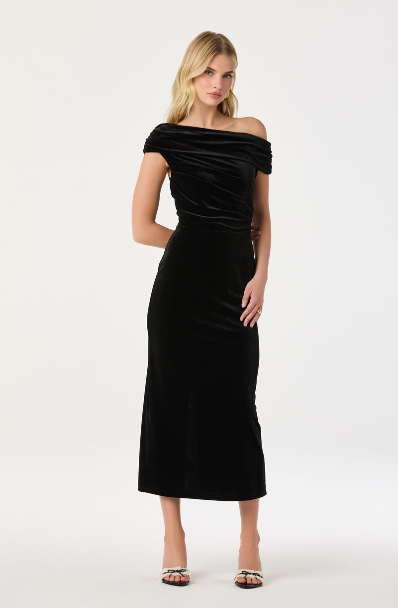 A woman is wearing a black off-the-shoulder dress made of a shiny, velvety fabric. The dress has a fitted bodice with draped detailing at the neckline and extends down to a midi length. She is also wearing open-toed high heels with a decorative element on the front.