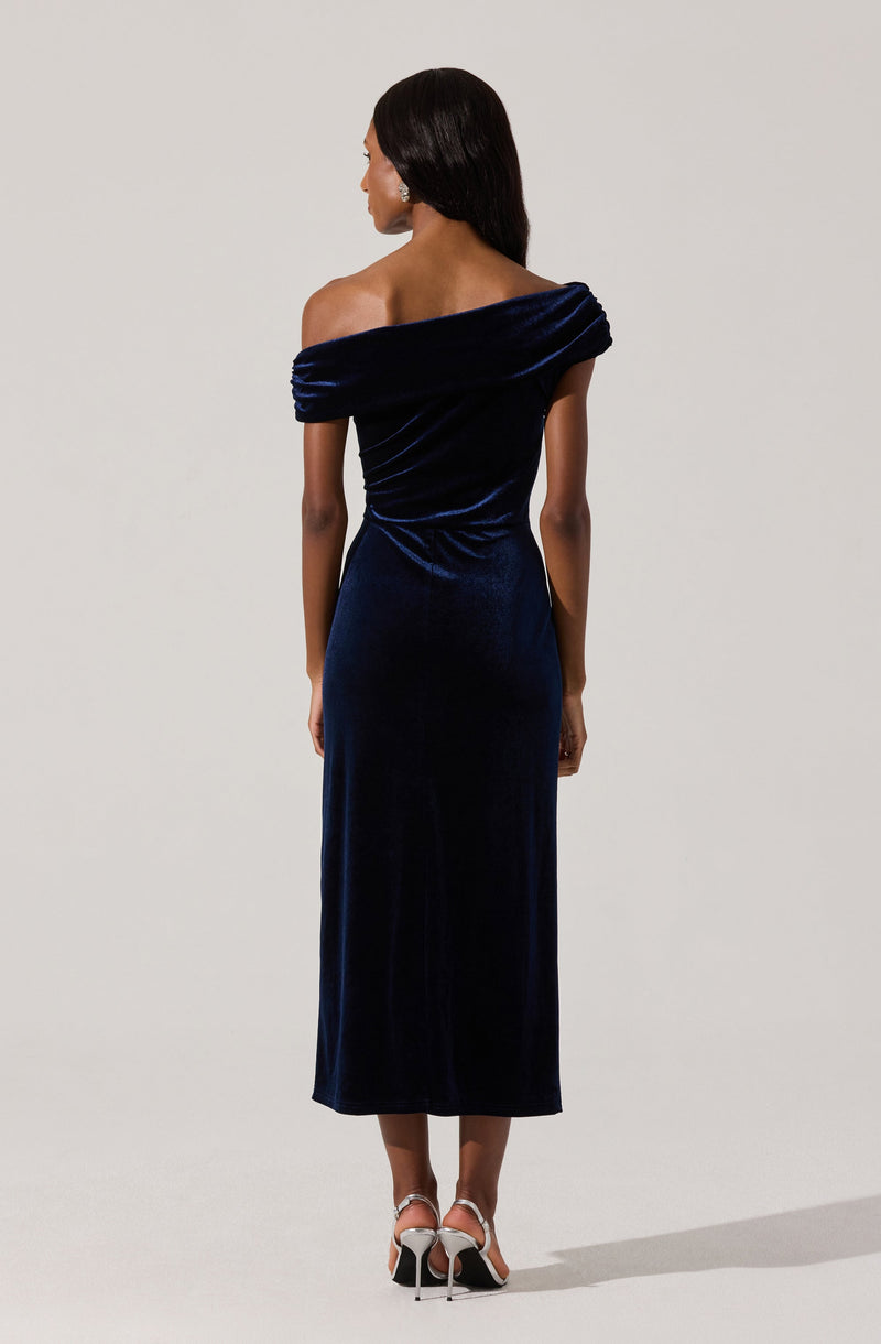 A woman is wearing a dark blue velvet off-the-shoulder gown with a draped neckline and long length. She is also wearing white high-heeled sandals.