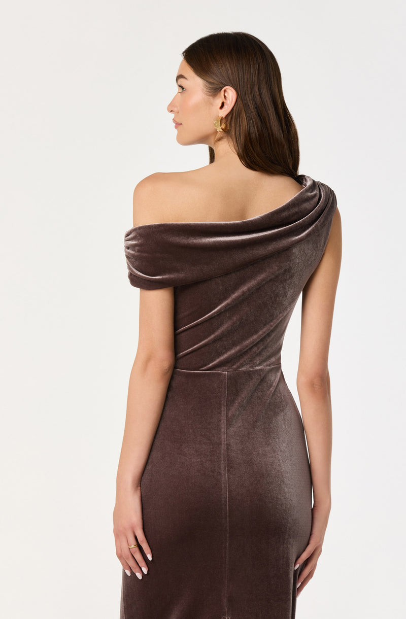 A woman is wearing a sleeveless, form-fitting, taupe velvet dress with an asymmetrical off-the-shoulder neckline that drapes over one shoulder.