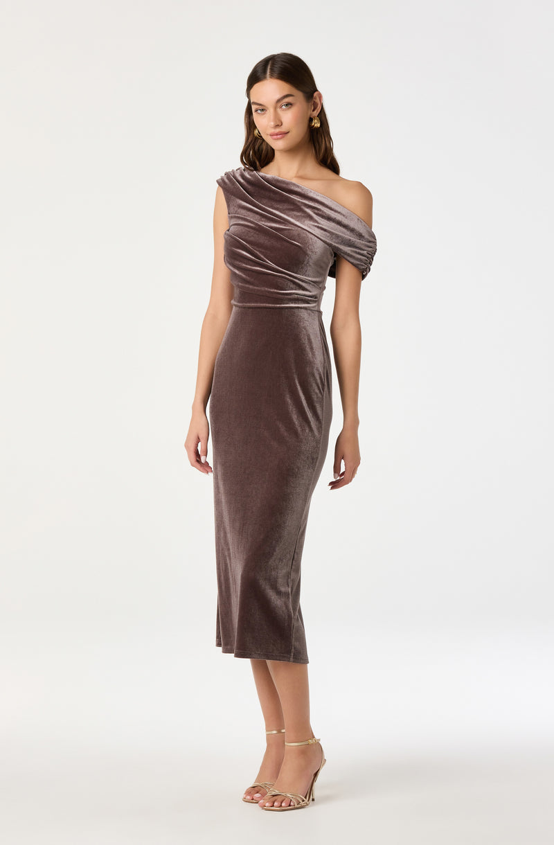 The dress is a form-fitting, mid-length gown made of soft, velvety fabric in a muted mauve color. It has an asymmetric, off-the-shoulder neckline with a gathered detail that drapes over one shoulder, creating a ruched effect across the bodice. The material has a smooth, shiny texture, giving it an elegant appearance. The dress tapers down to the calves, emphasizing a sleek silhouette.