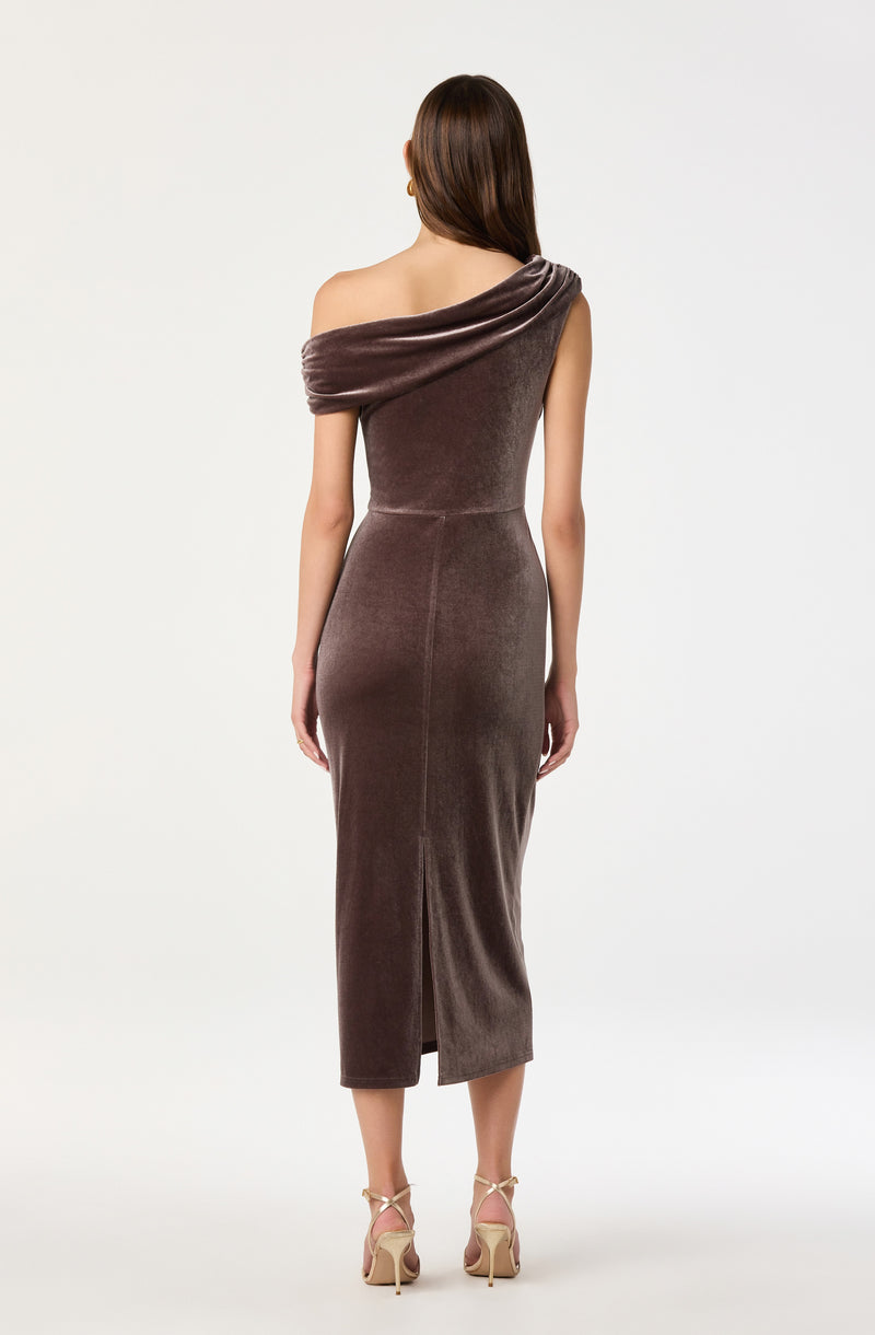 The woman is wearing an elegant, floor-length dress made of a shiny, velvety fabric in a taupe or brown color. The dress features a one-shoulder design with a draped, wide strap that wraps around her shoulder, creating a sophisticated look. She is also wearing nude-colored high-heeled sandals with ankle straps.