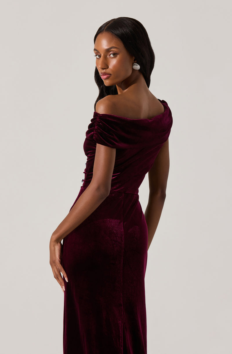 A woman wearing an off-the-shoulder dress made of deep burgundy velvet fabric. The dress features ruched detailing on the shoulders and a fitted silhouette that extends down to the floor.