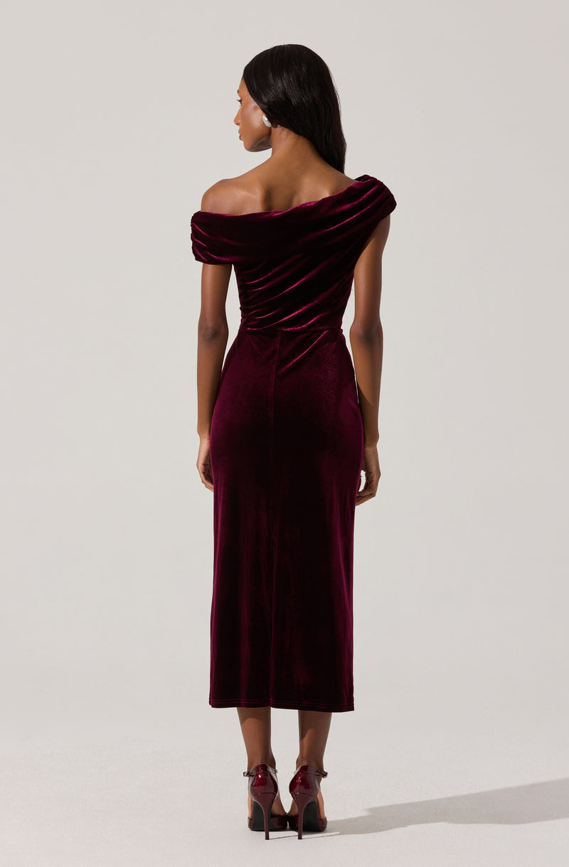 A woman is wearing an off-the-shoulder, deep burgundy velvet dress that extends to mid-calf length. The dress features a gathered neckline draping over her shoulders and a form-fitting silhouette. She is also wearing matching burgundy high heels with ankle straps.