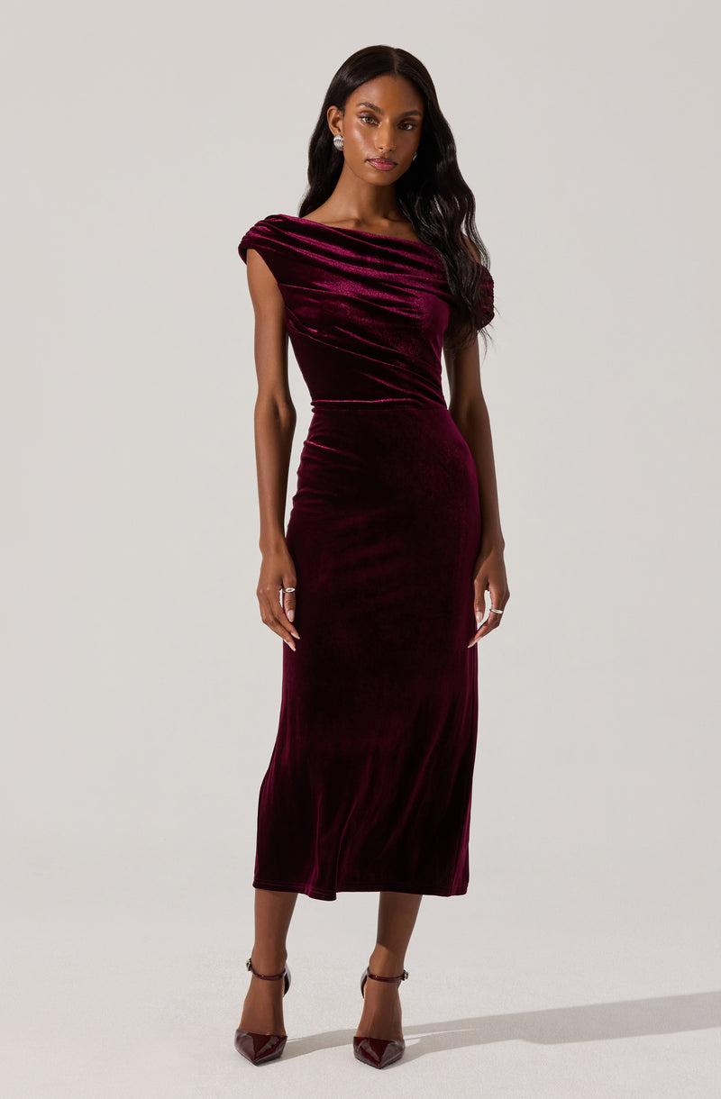 A woman is wearing a sleeveless, floor-length velvet dress in a deep burgundy color. The dress features a fitted silhouette with ruching at the bodice and a subtle draped detail at the shoulder. She completes the look with matching burgundy high-heeled shoes.
