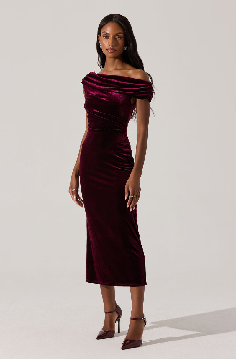 A woman is wearing an elegant, off-the-shoulder gown made of deep burgundy velvet. The dress features a ruffled neckline that drapes over her shoulders and a fitted silhouette that extends to a midi length, reaching just above her ankles. She pairs the dress with matching burgundy high-heeled shoes.