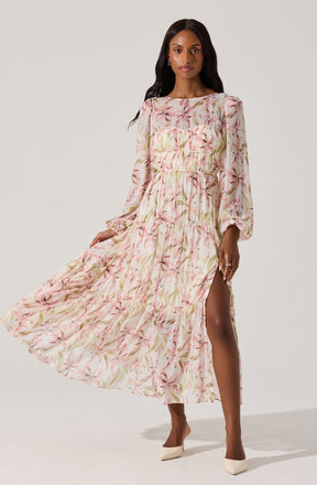 A woman is wearing a long, flowing dress with a light, pastel-colored floral print. The dress features long sleeves with elastic cuffs, a round neckline, and a tiered, ruffled skirt with a high slit on one side. The fabric appears lightweight and semi-sheer, with a soft pink and beige color palette accented by green leaves and pinkish flowers. She pairs the dress with beige high heels.