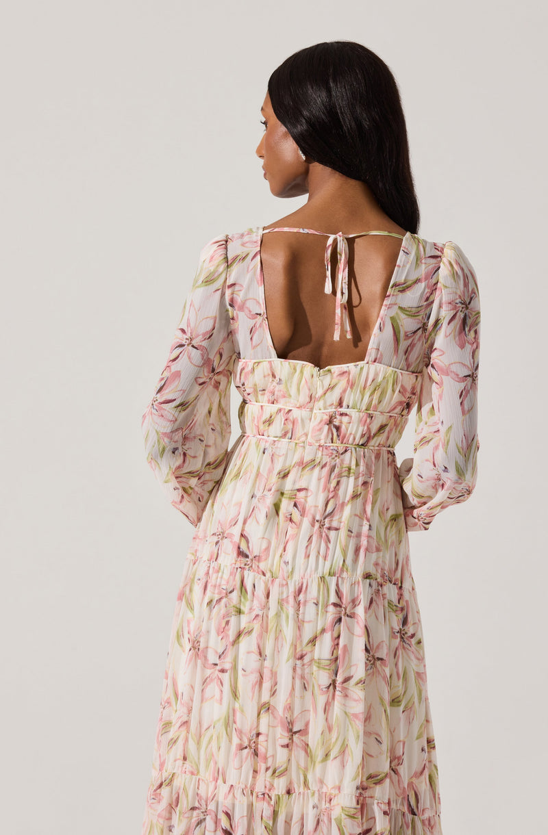 A woman is wearing a long, flowy dress with a floral pattern in shades of pink, green, and cream. The dress features puffed long sleeves and a square neckline with a keyhole opening at the back, tied with a thin ribbon. The fabric appears to be lightweight and slightly sheer.