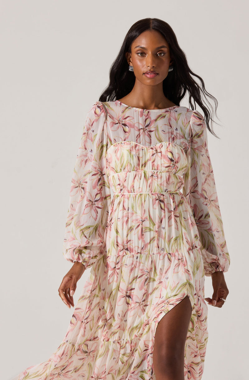 A woman is wearing a long, flowing dress with a floral pattern that features pink, green, and beige colors. The dress has long puffed sleeves with elastic cuffs and a ruffled slit that reveals her leg. The fabric appears lightweight and semi-sheer, with a gathered and tiered design creating a textured look.