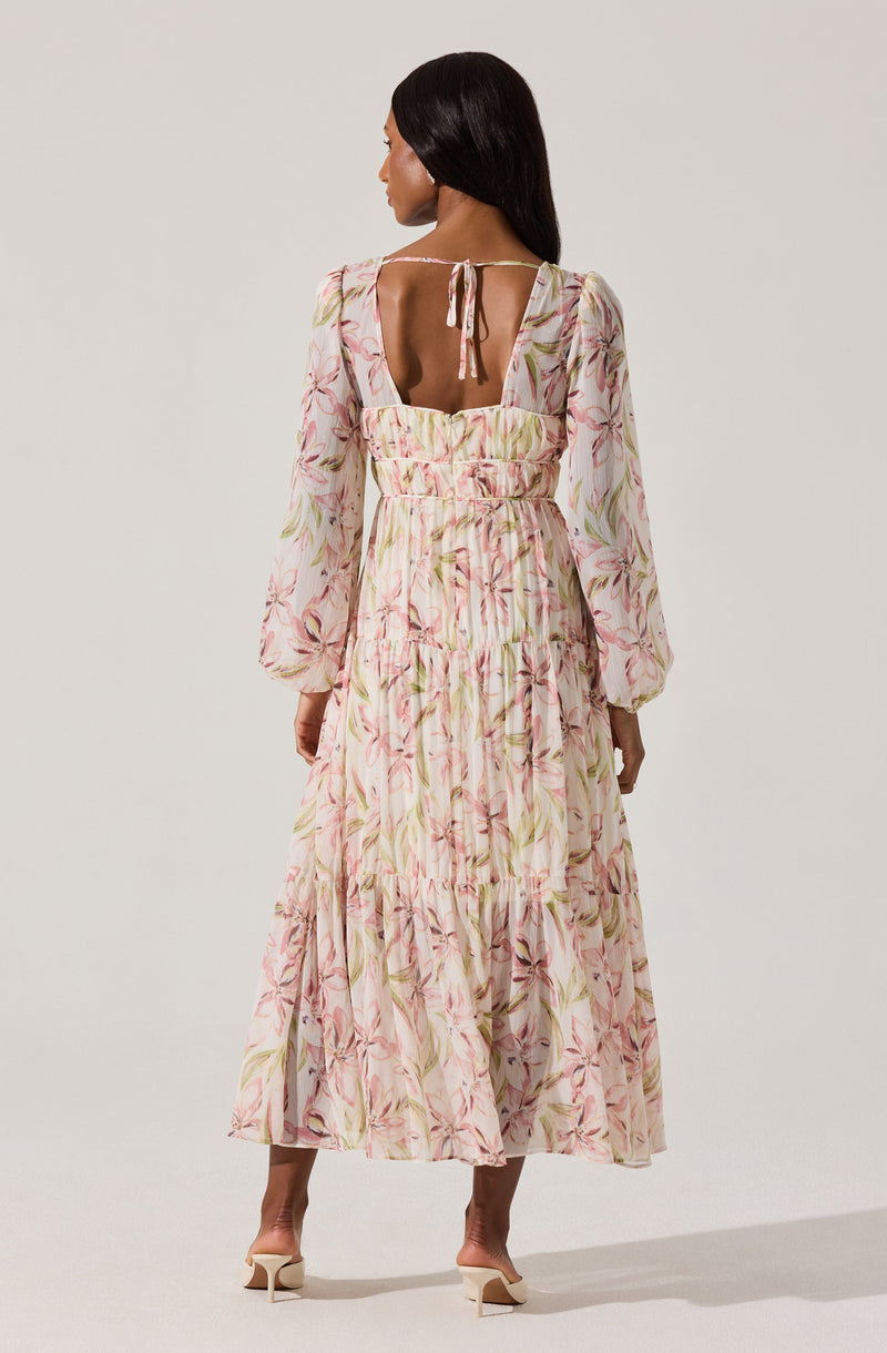 A woman is wearing a long, flowy dress with a light background and a soft pink, green, and beige floral pattern. The dress features puffy long sleeves, a fitted waist, and a tiered skirt. The back of the dress has a square cutout with a small bow tied at the top of the opening.