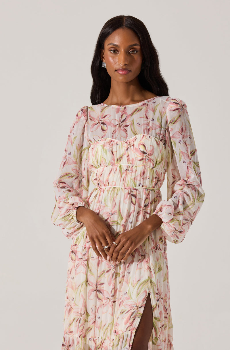 A woman is wearing a long, flowing dress with a pastel floral pattern. The dress has long, puffed sleeves that are gathered at the cuffs, and a high slit on one side revealing her leg. The fabric features soft pink and green flowers with light-colored leaves.