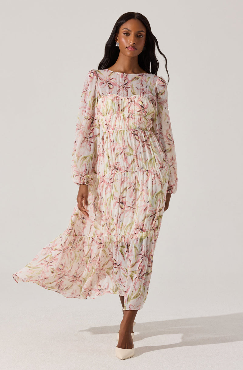 A woman is wearing a long, flowing dress with a pastel floral print featuring pink, green, and beige tones. The dress has long, sheer sleeves with elastic cuffs and a round neckline. The fabric is gathered at the waist, creating a tiered, ruffled effect throughout the dress. She pairs the outfit with white pointed-toe shoes.